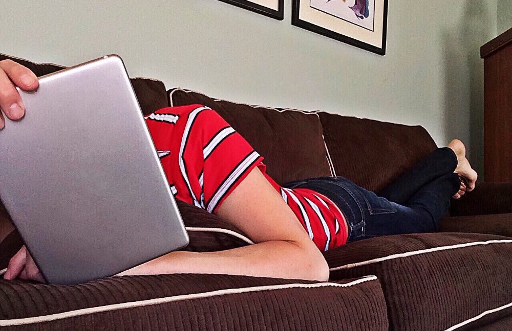 Person lying on a brown couch covering their head with a tablet