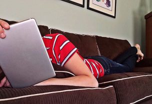 Person lying on a brown couch covering their head with a tablet