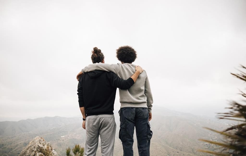 Two men holding hands over shoulders looking into the distance from atop a hill